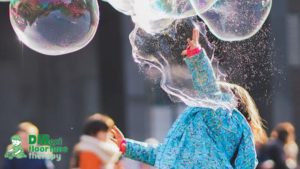 A young girl in a blue sweatshirt bursts bubbles that float above her, with her face hidden from view.