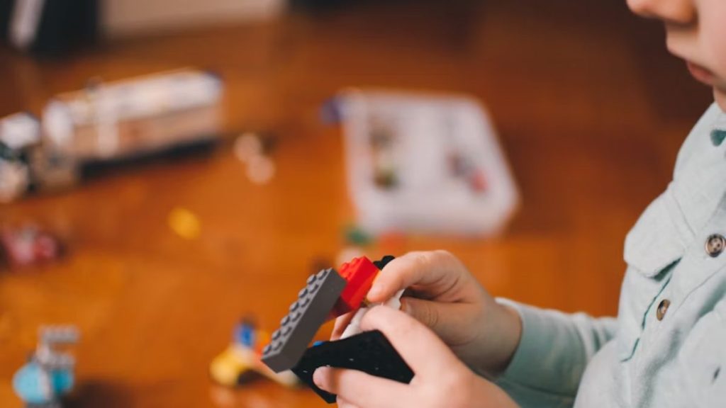 A young boy plays with colored blocks in a quiet room, with only his hands and profile visible, appearing to be alone.