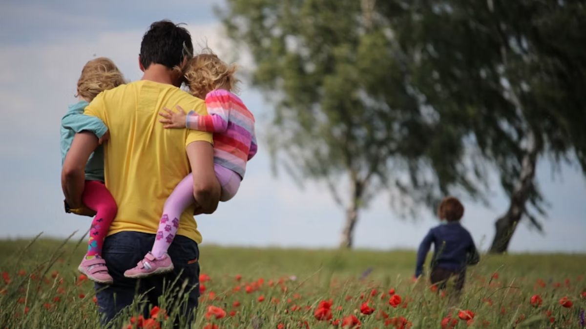 A father carries his two children, one on each arm, walking through a field of colorful red flowers and a wide expanse of grass.