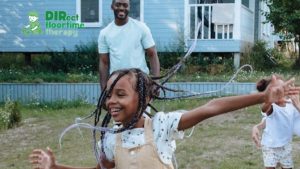 A young girl happily plays on a lawn outside her home while her father watches over her.
