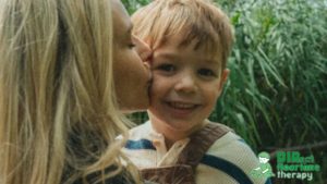 A blonde mother kisses her young blonde son in the garden, surrounded by greenery, sharing a tender bonding moment.