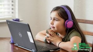 A young girl wearing pink headphones looks at a laptop, appearing relaxed and focused, dressed in a casual shirt.