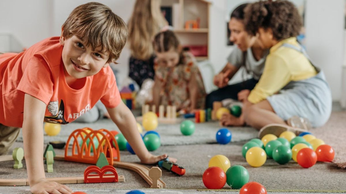 A boy gazes at the camera while other children play in the background, building with bricks and discs, softly out of focus.