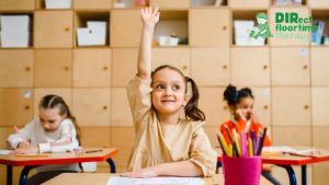 A young girl raises her hand in the classroom, looking confident and energetic, while other children write in their notebooks in the background.
