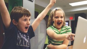 A boy and a girl smiling triumphantly while looking at a computer monitor, proud of their school performance in a colored photo.