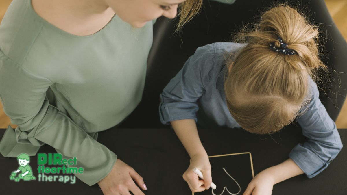 A young girl draws a heart on a chalkboard while a female therapist looks on and guides her during a DIR/Floortime session.