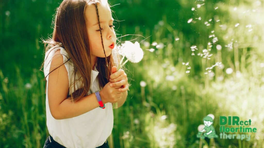 A young child blows on dandelion wishing flowers, exploring and imagining through the DIR Floortime model.