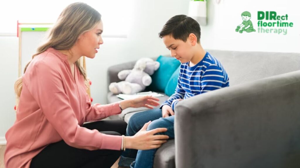 Female therapist talking with a young boy in a blue shirt on the couch during behavioral therapy for ADHD; the child looks serious and attentive.