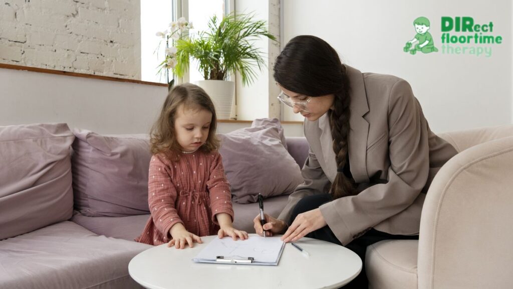Female therapist leaning on a table with a young girl in a flower-printed dress, both looking at records; the child appears serious and focused during behavioral therapy for ADHD.