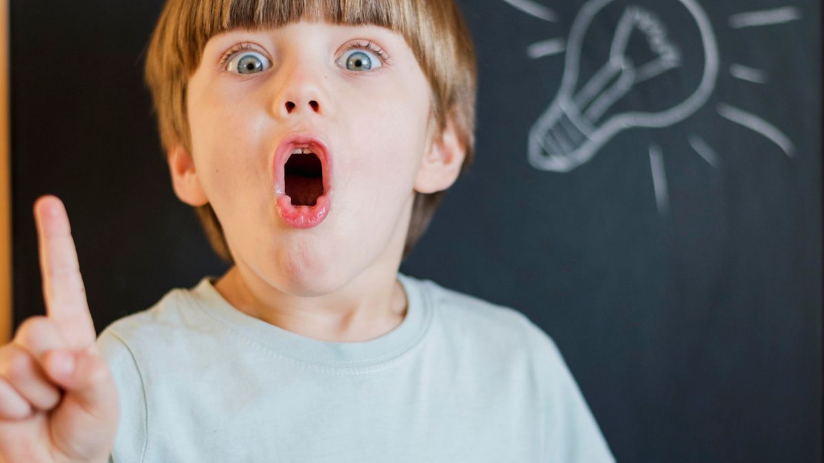 A young boy with a surprised expression raises his finger, standing in front of a board with a lightbulb illustration, representing brain games for kids.