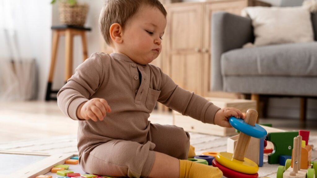 A 6-month-old, dressed in a chocolate-colored onesie, carefully arranges puzzle pieces across the soft carpeted floor, exploring shapes and textures.