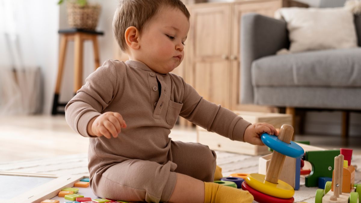 A 6-month-old, dressed in a chocolate-colored onesie, carefully arranges puzzle pieces across the soft carpeted floor, exploring shapes and textures.
