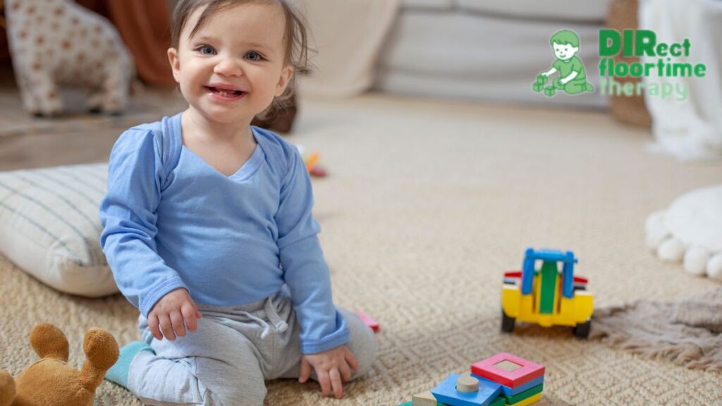 A toddler in a cornflower blue shirt sits confidently on the floor, smiling at the camera with toys arranged nearby.