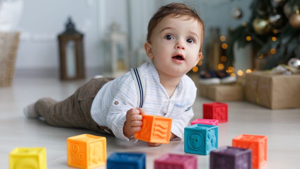 A toddler in a white shirt and brown suspenders plays tummy time on the floor with colorful blocks, exploring and reaching for the toys.