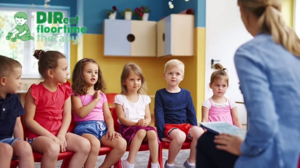 A group of children sit side by side, engaging with a female therapist whose back is to the camera and slightly blurred, during a DIR Floortime speech therapy session.