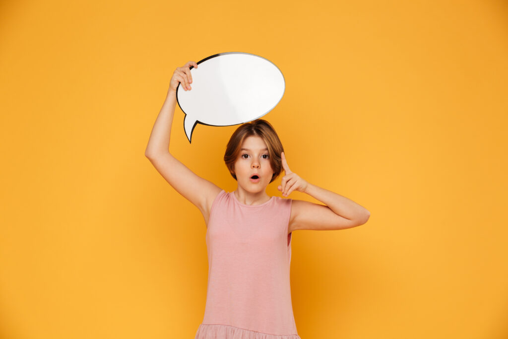 A young girl in a pink shirt holds up a speech bubble above her head and flashes a thumbs-up during a DIR Floortime speech therapy session.