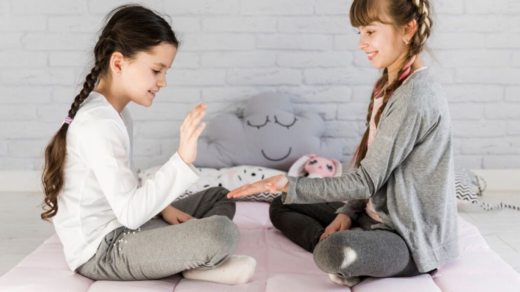 Two young girls play hand and puzzle games on a beige rug on the floor.