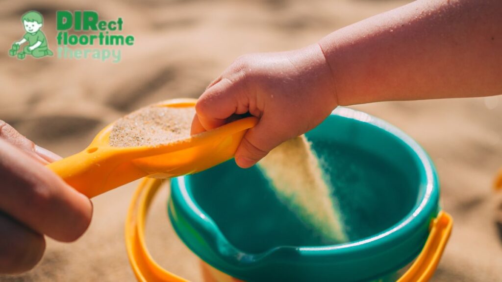 Two toddlers’ hands pour sand into a small bucket as part of a sensory bin activity.