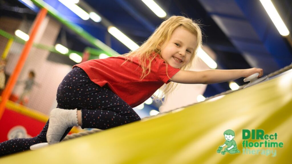 A young blonde girl climbs and slides down an obstacle course during movement play.