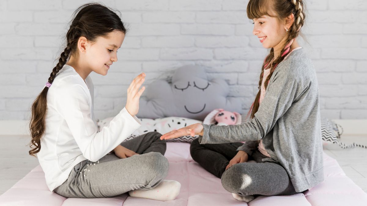 Two young girls play hand and puzzle games on a beige rug on the floor.