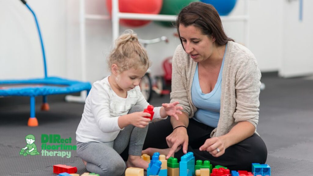 A female therapist and a young girl engage in a structured play therapy session on a gray foam-padded floor. The young girl, with blonde hair tied in a high bun and wearing a white long-sleeve shirt and gray leggings, carefully manipulates a large red building block. The therapist, wearing a beige cardigan over a light blue top, sits close beside her, leaning forward with an encouraging expression and pointing toward a row of blue and green interlocking blocks. A blue mini-trampoline and large exercise balls are visible in the background, and the green "DIRect floortime therapy" logo is placed in the bottom-left corner.