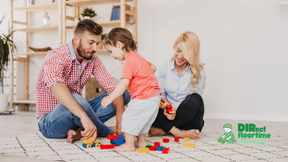 A mother, father, and young child participate in a DIRFloortime therapy session at home on a patterned area rug. The father, wearing a red-and-white checkered shirt and jeans, sits cross-legged and playfully interacts with a toy truck. The young child, dressed in an orange t-shirt and light shorts, stands bare-footed, focusing intently on stacking colorful toy blocks on the floor. The smiling mother sits cross-legged nearby, holding red blocks and warmly observing the interaction. In the bottom-right corner, a green logo reads "DIRect floortime therapy