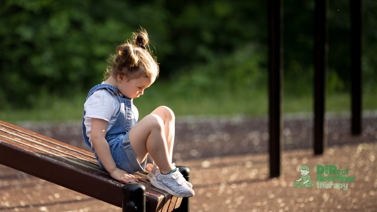 A girl sitting slightly apart from a group of children at a playground, observing carefully rather than joining, looking thoughtful rather than distressed