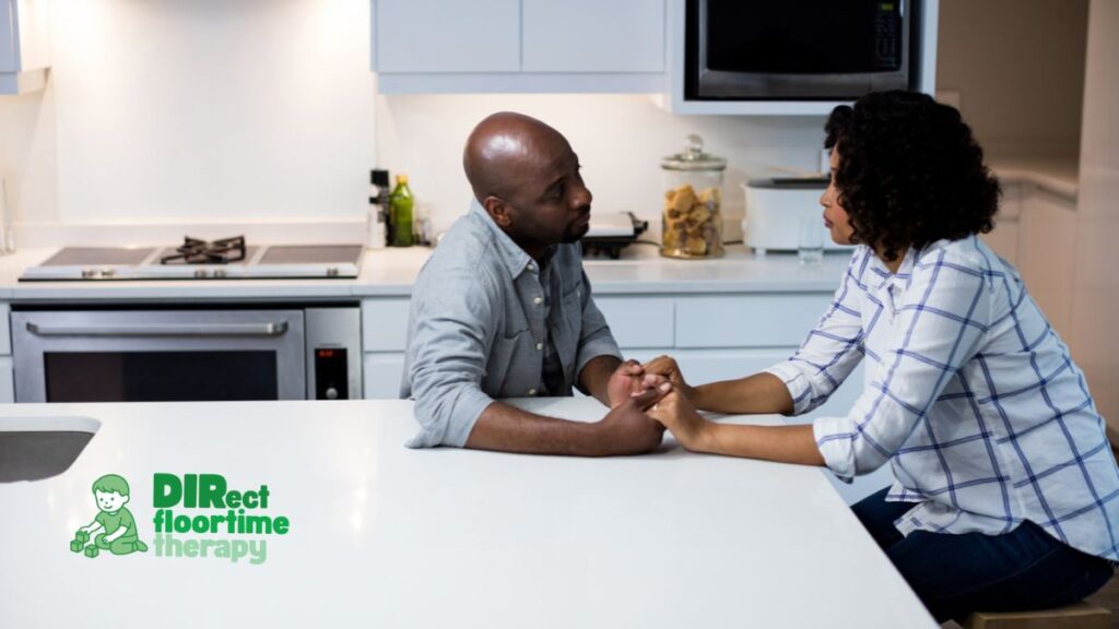 "Tired couple sitting across from each other at a kitchen island holding hands, supporting their relationship and co-regulation while raising an autistic child.