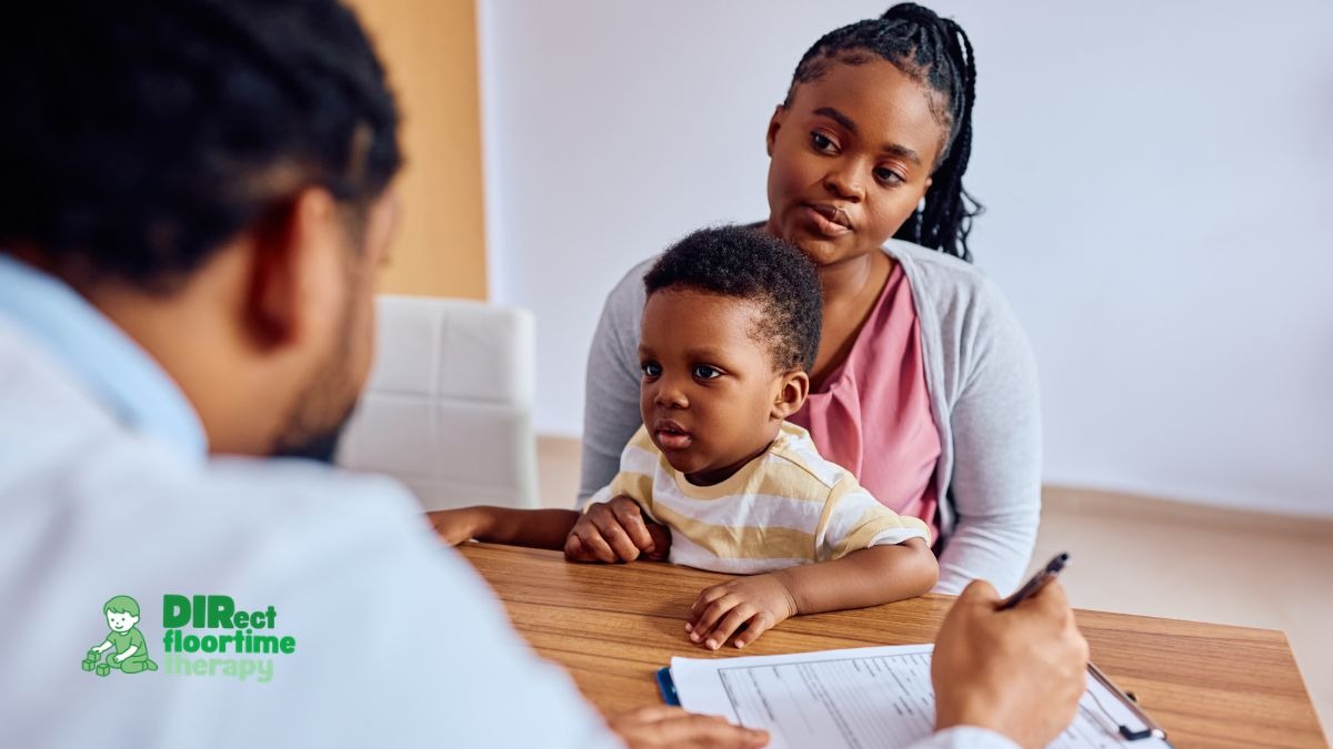 : A mother sitting at a wooden desk holding her young child while speaking to a healthcare professional, seen in foreground over the shoulder, writing on a clipboard, with the Direct Floortime Therapy logo in the bottom-left corner.