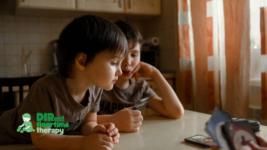 Two young boys, an autistic child and his sibling, sitting closely together at a table focused on playing a card game during a DIR Floortime activity.