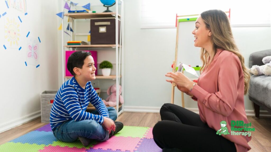 How to Talk to Your Child's School About DIR/Floortime and IEP Supports 1 A teacher or therapist sitting on the floor with a young autistic child, both engaged with a toy or activity. The adult is at the child's level, smiling, following the child's lead. Bright, warm classroom or therapy room setting.