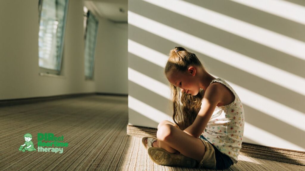 A child sitting quietly with their head down in a calm, low-lit room while an adult sits nearby, present but not intrusive. Conveys the emotional contrast between internal distress and outward stillness. Warm, soft tones. No text overlay.